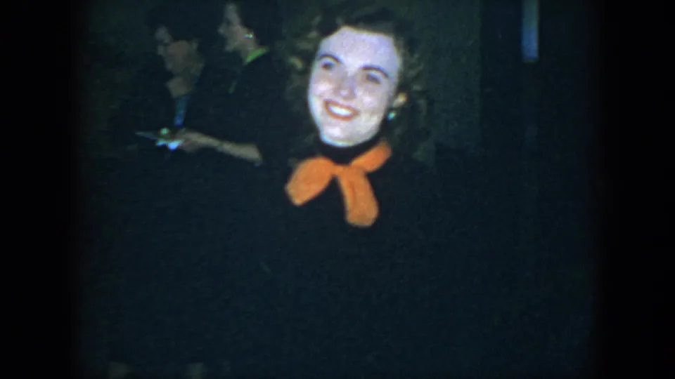 A close 1952 Houston home-movie frame shows the woman smiling and tilting her head, with a dim background guest holding a drink.