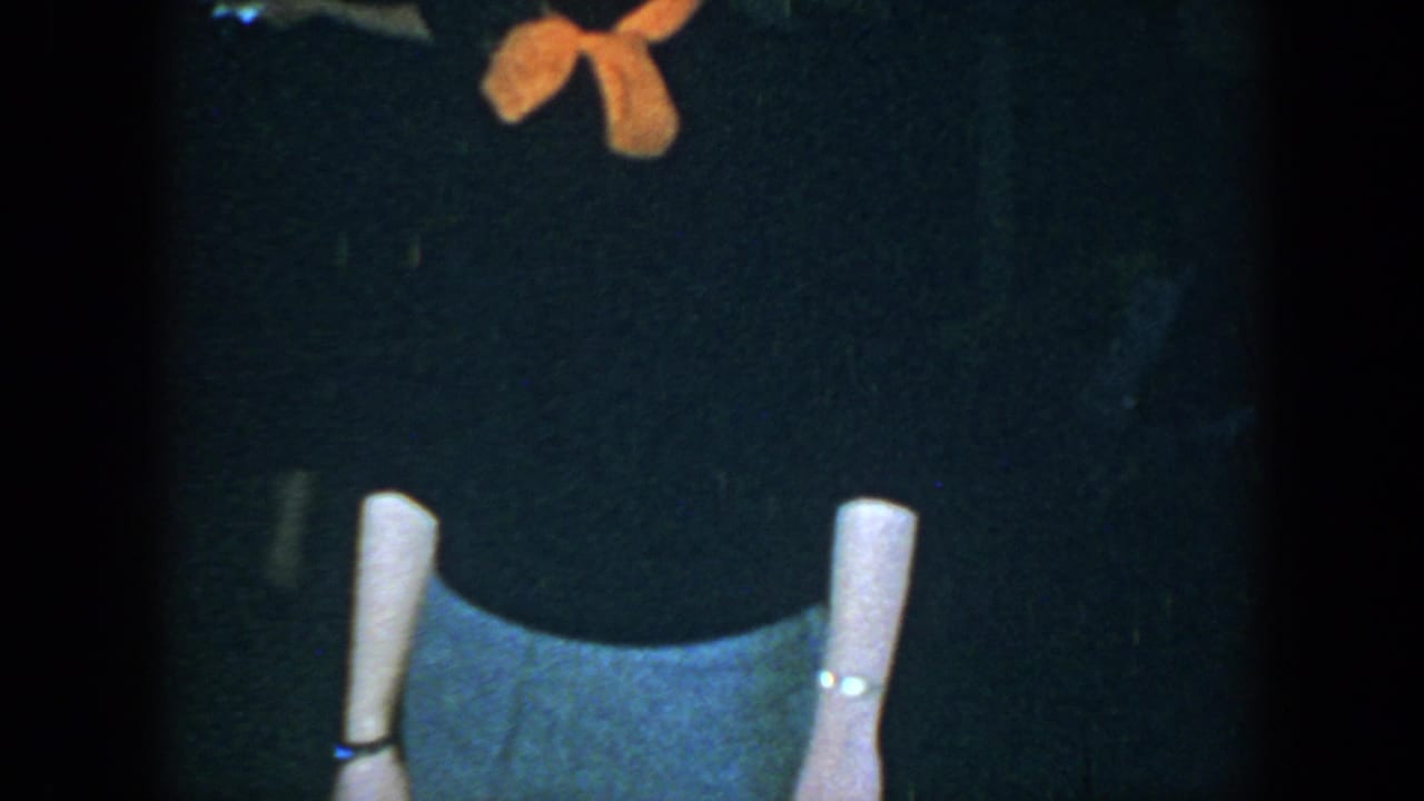 Adult woman in a dark top, orange scarf, gray skirt, white socks, and dark shoes posing against a dark background.