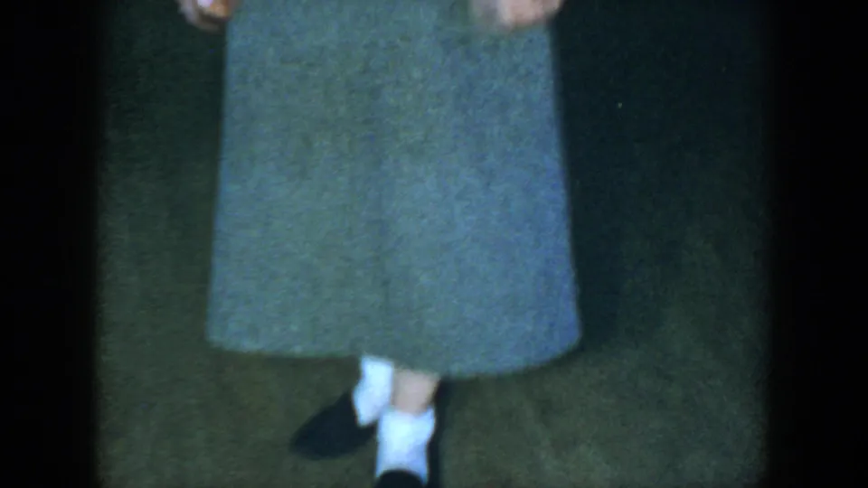 The home-movie frame centers on the woman's gray skirt, white socks, and dark shoes, with her upper body partly cropped.