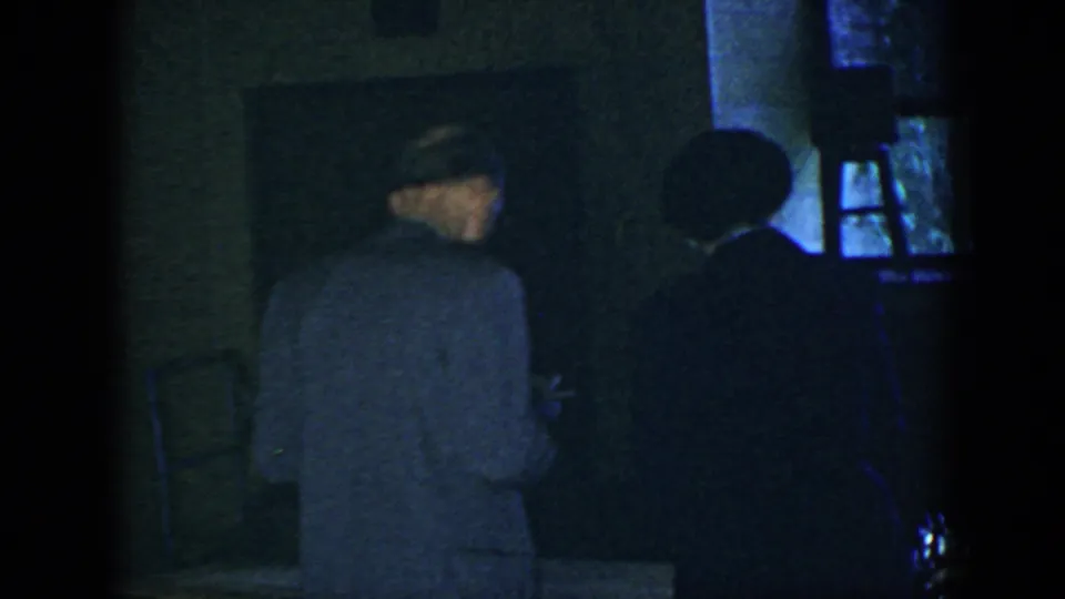 1952 Houston home-movie frame showing adults gathered in a dark private interior, with one man turned away near the windows.