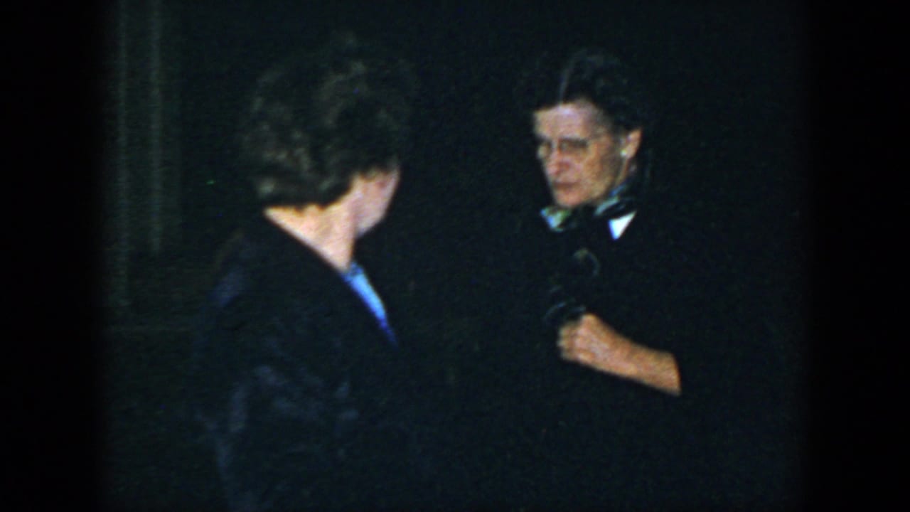 Two adults in dark clothing face each other in a dim room.