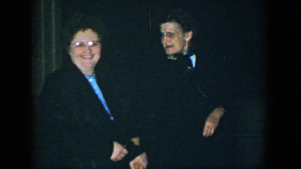 A woman with glasses smiles beside a man in a bow tie during a dim 1952 Houston indoor gathering.