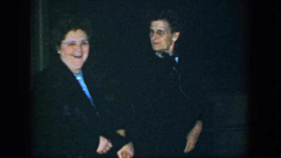 The woman smiles toward the camera as the man stands slightly behind her in dim 1952 Houston party footage.