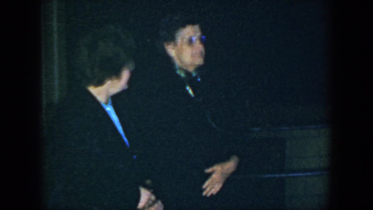 Two women in dark coats stand close together in a dim nighttime setting.
