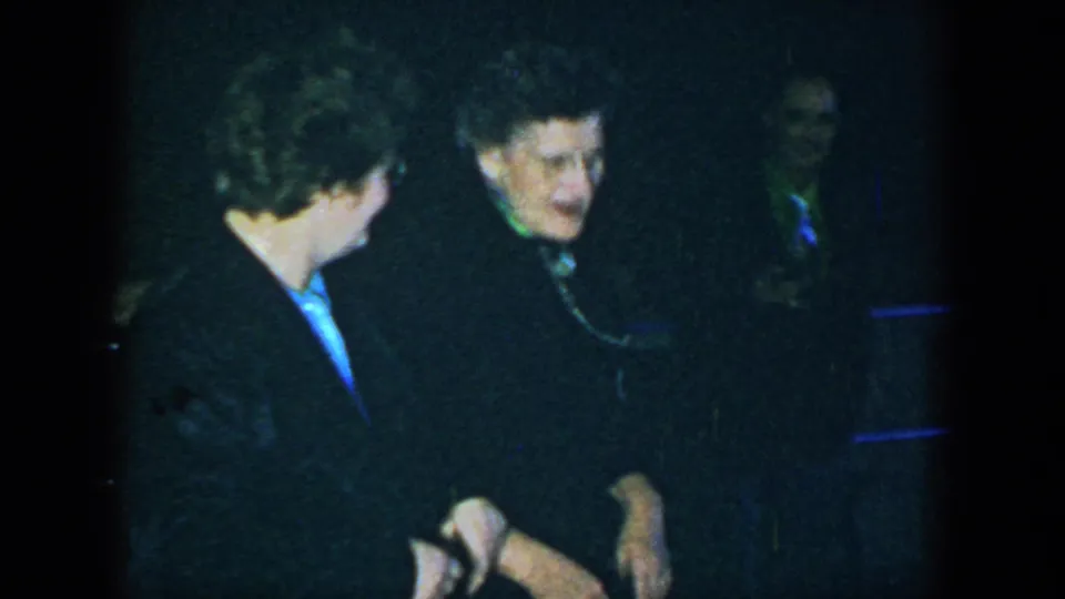 Two women in dark coats converse at night in Houston, with a third figure partly visible behind them.