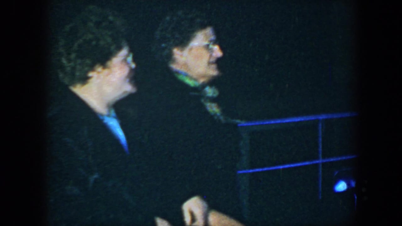 Two women in dark clothing stand in a dim interior, one wearing glasses and a green scarf.