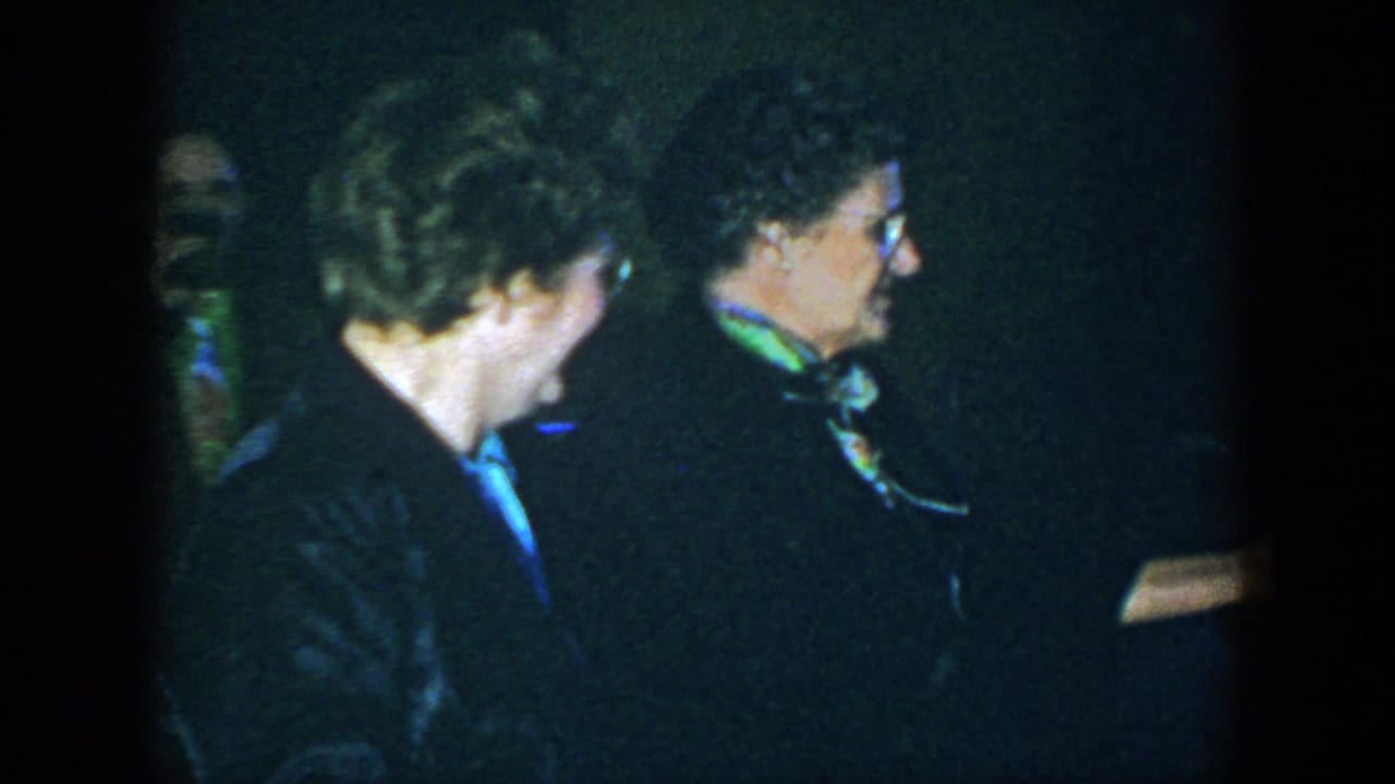 Two women are seen in profile in a dim interior, including one with glasses and a green scarf.