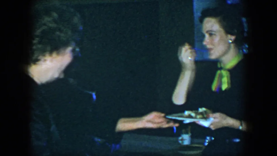A woman in a dark dress with a bright green neck scarf holds a plate of food at the Houston social gathering.