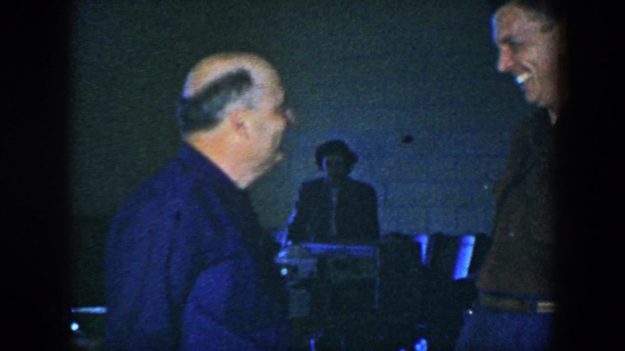 Two adult men laugh in a dim room with a white brick wall and chairs behind them.