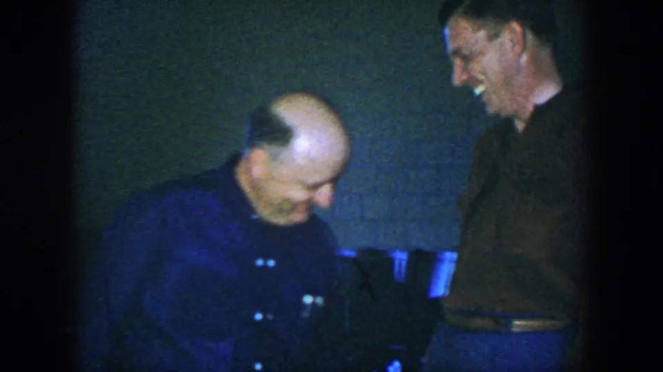 A balding man in a dark shirt leans forward laughing as a second adult man smiles in the dim 1952 Houston interior.