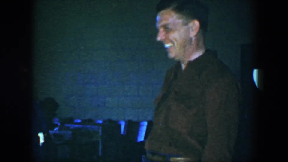 A smiling adult man in a brown shirt stands in front of chairs, with a partially obscured person in the dim Houston interior.