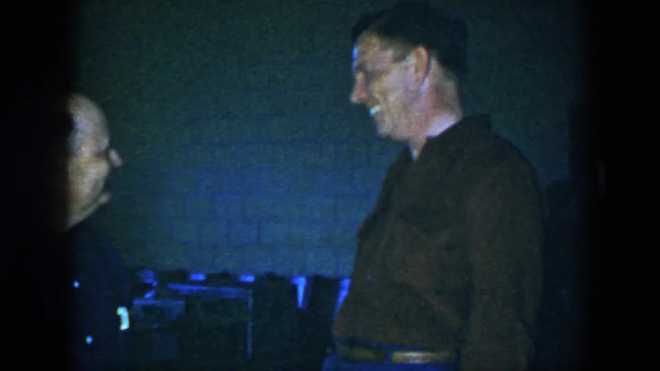 The 1952 Houston home-movie frame shows a smiling adult man in a brown shirt facing left against a dim white brick interior.