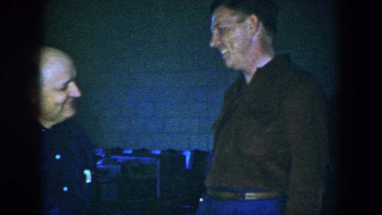 In 1952 Houston home-movie footage, two adult men face each other in a dim interior near a blue-tinted brick wall and seating.