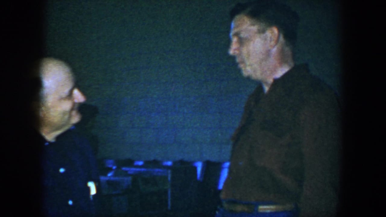 A seated or shorter man looks toward a taller man standing beside a brick wall.
