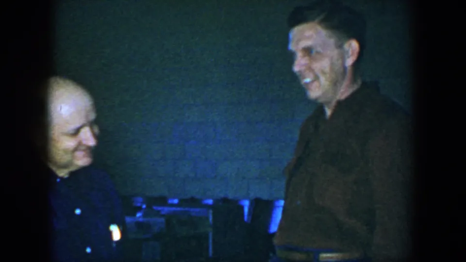 A smiling exchange continues in the 1952 Houston interior, with blue-green 8mm color shift and a brick wall in the background.