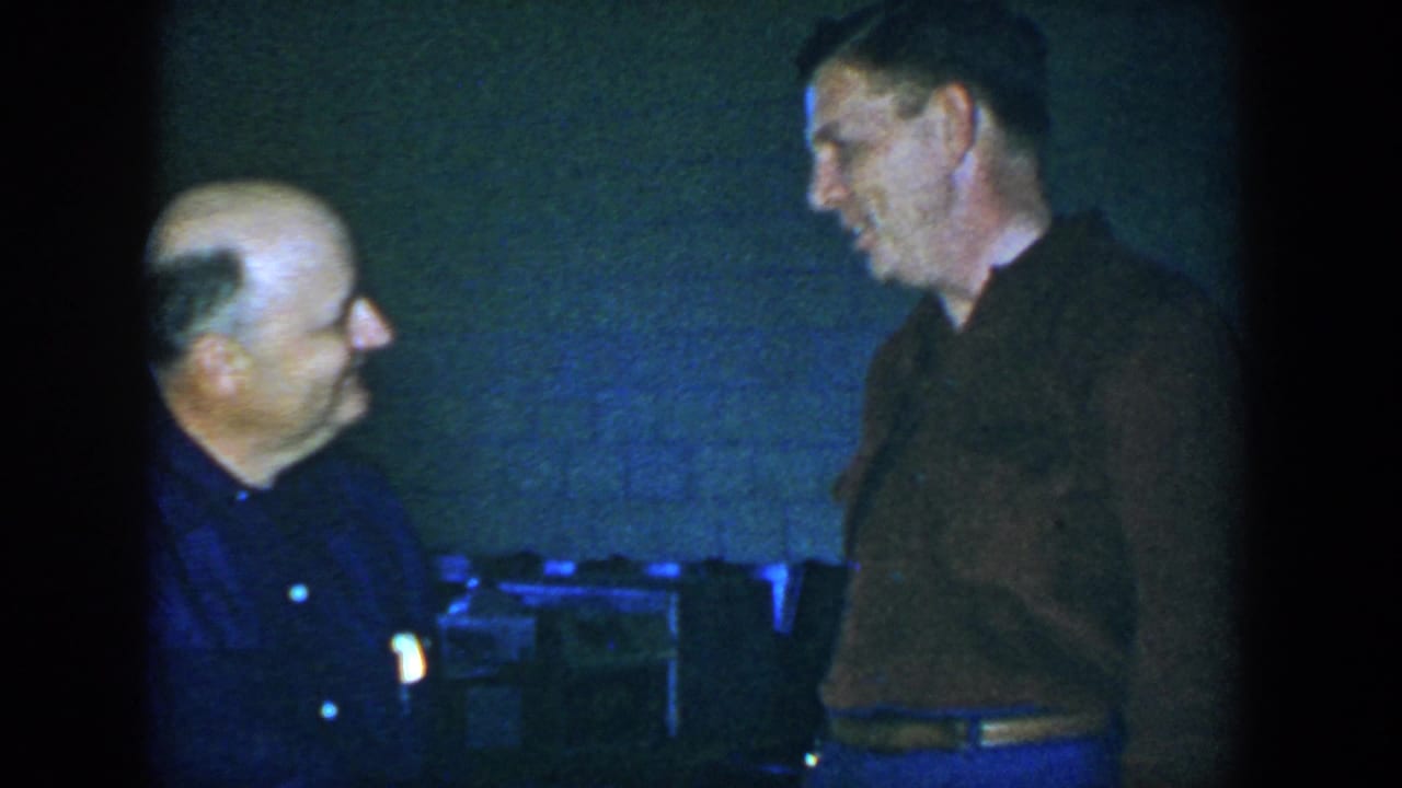 Two adult men face each other in a dim room, one seated and one standing.