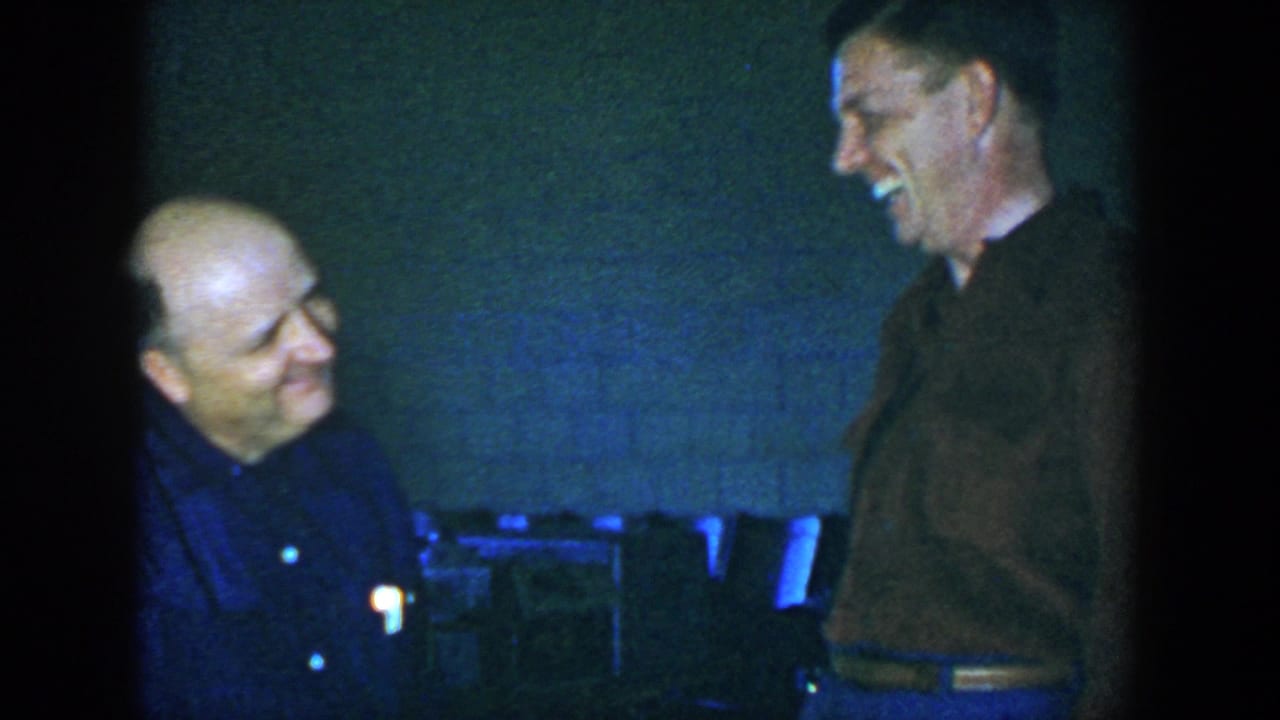 The two men smile while talking in the same dim interior.