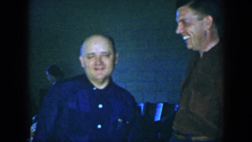 The seated balding man turns toward the camera while the standing man laughs, filmed in a dim Houston interior in 1952.