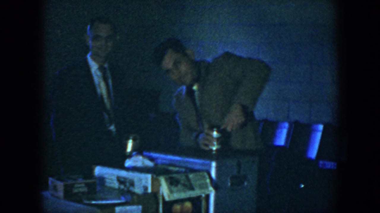Two suited men stand near a table with a box in a dim room.