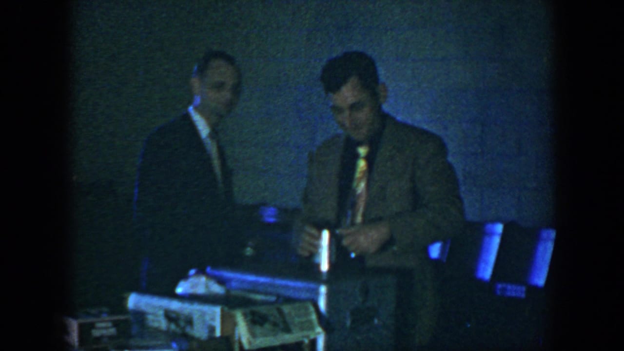 In the dim 1952 Houston interior, a suited man appears to handle a small object near a case while another man observes.