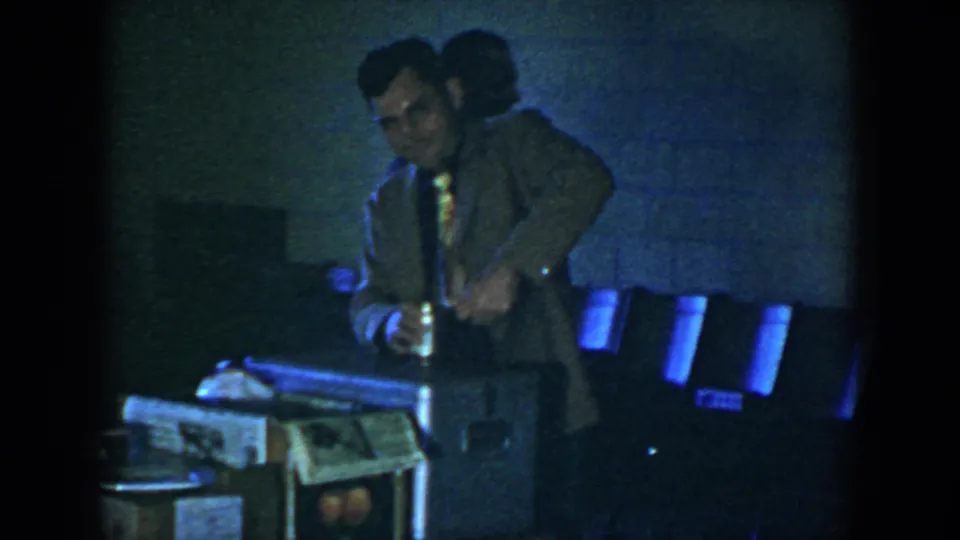 A 1952 Houston home-movie frame shows a man in a light suit and patterned tie handling an item beside a case on a table.