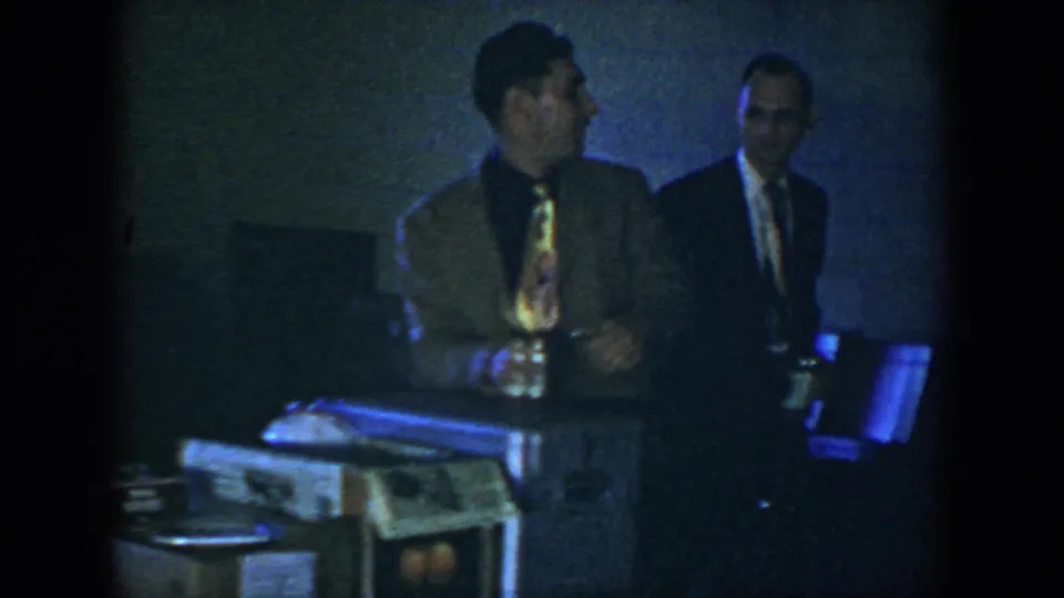 Near the end of the 1952 Houston clip, two suited men stand by the table as the man in front turns while holding a small object.