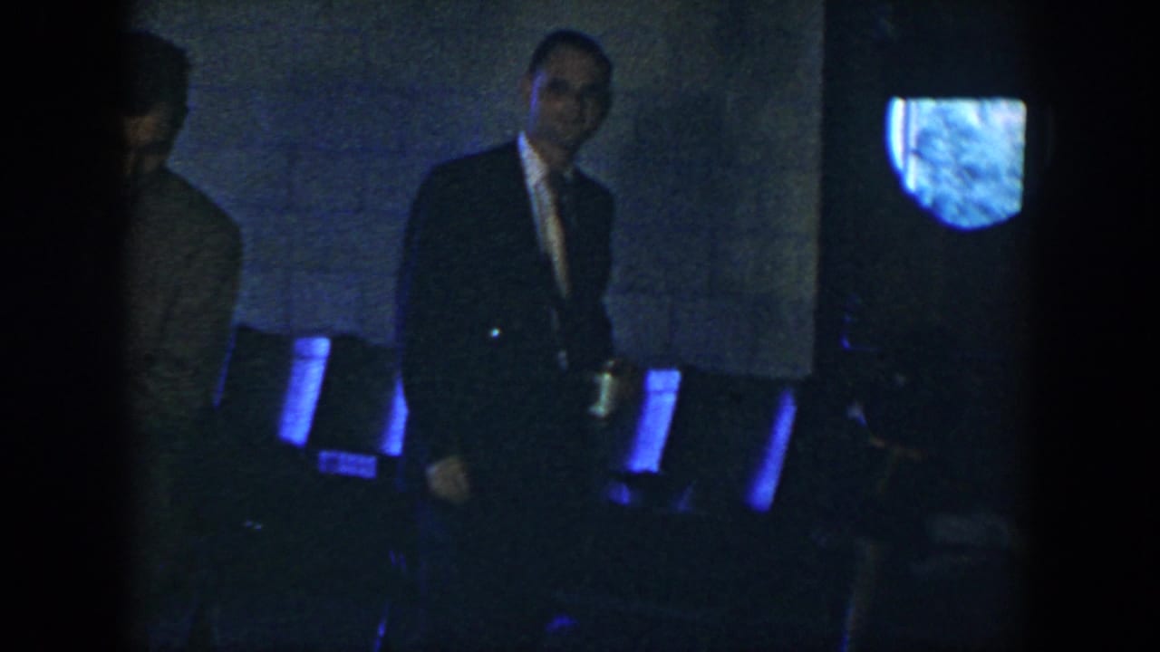 A suited man holding a can stands in a dark Houston interior, with chairs and a partly obscured adult figure behind him.