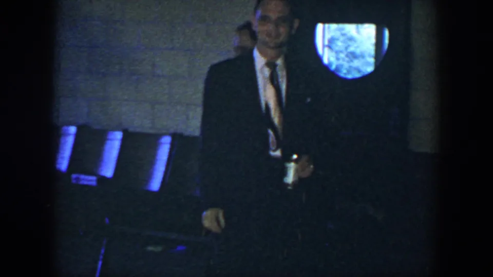 A suited man in the 1952 Houston clip holds a can while moving near chairs and a blue-lit opening in the wall.