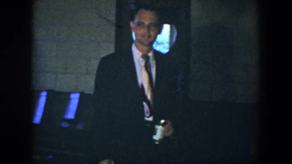 A suited man faces the camera in the Houston home-movie scene, holding a beer can near chairs and a blue-lit opening.
