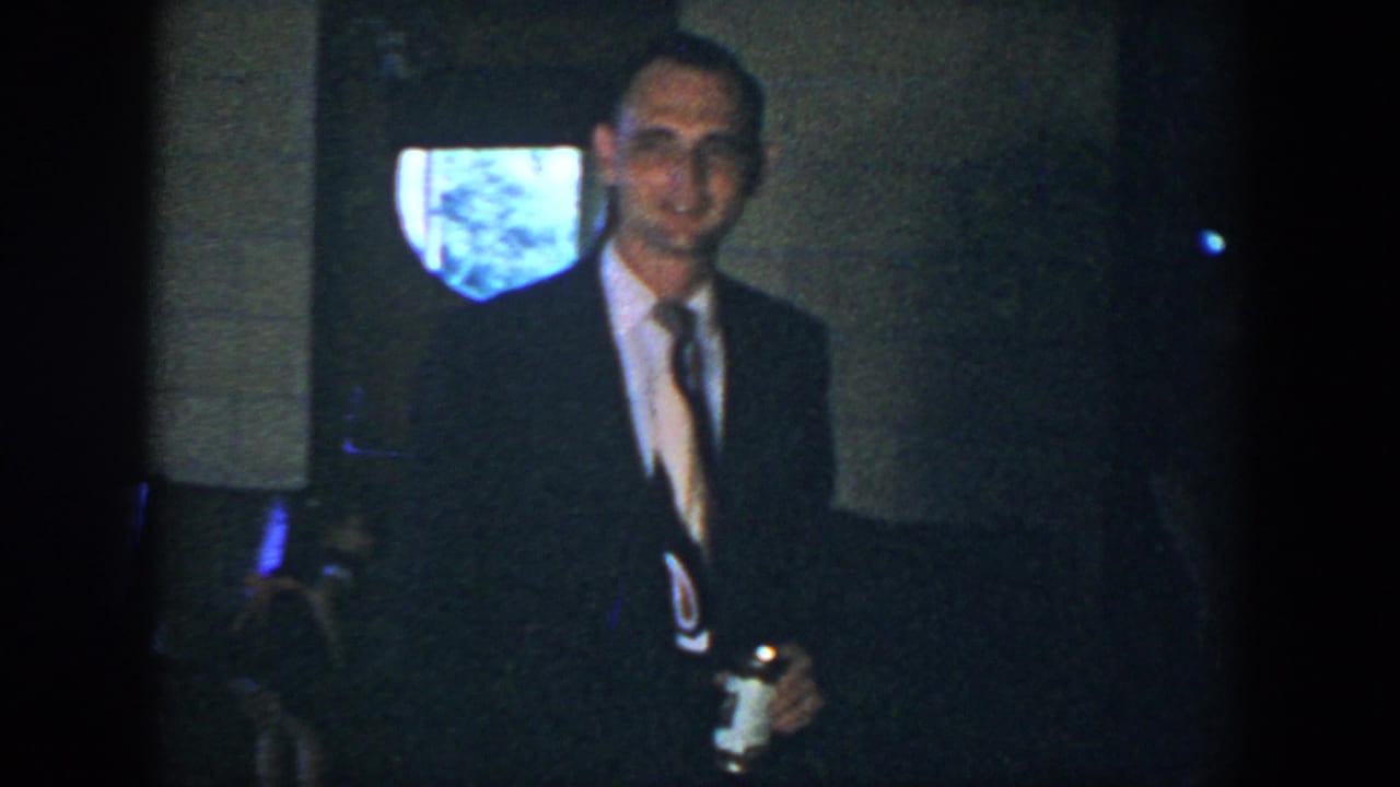 Man in a dark suit and tie smiles indoors while holding a drink can.
