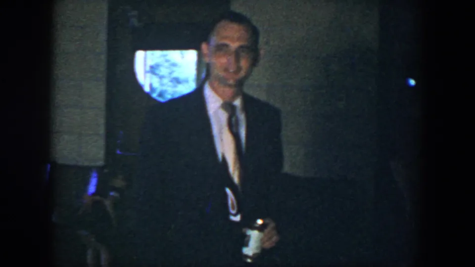 In supported 1952 Houston home-movie footage, a man in a dark suit and patterned tie smiles in a dim interior while holding a can.