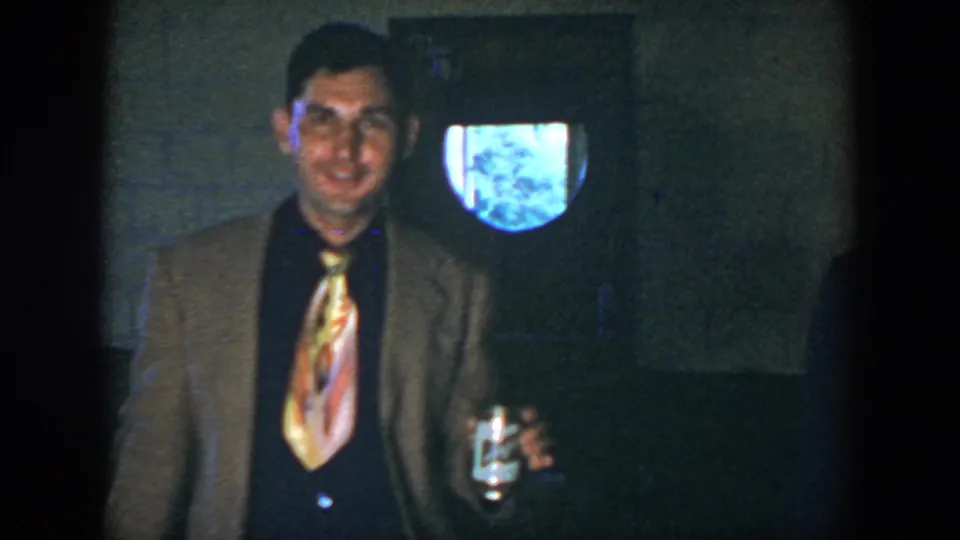 The man in the brown jacket smiles toward the camera in the supported 1952 Houston home-movie scene, holding a can beside a bright wall opening.