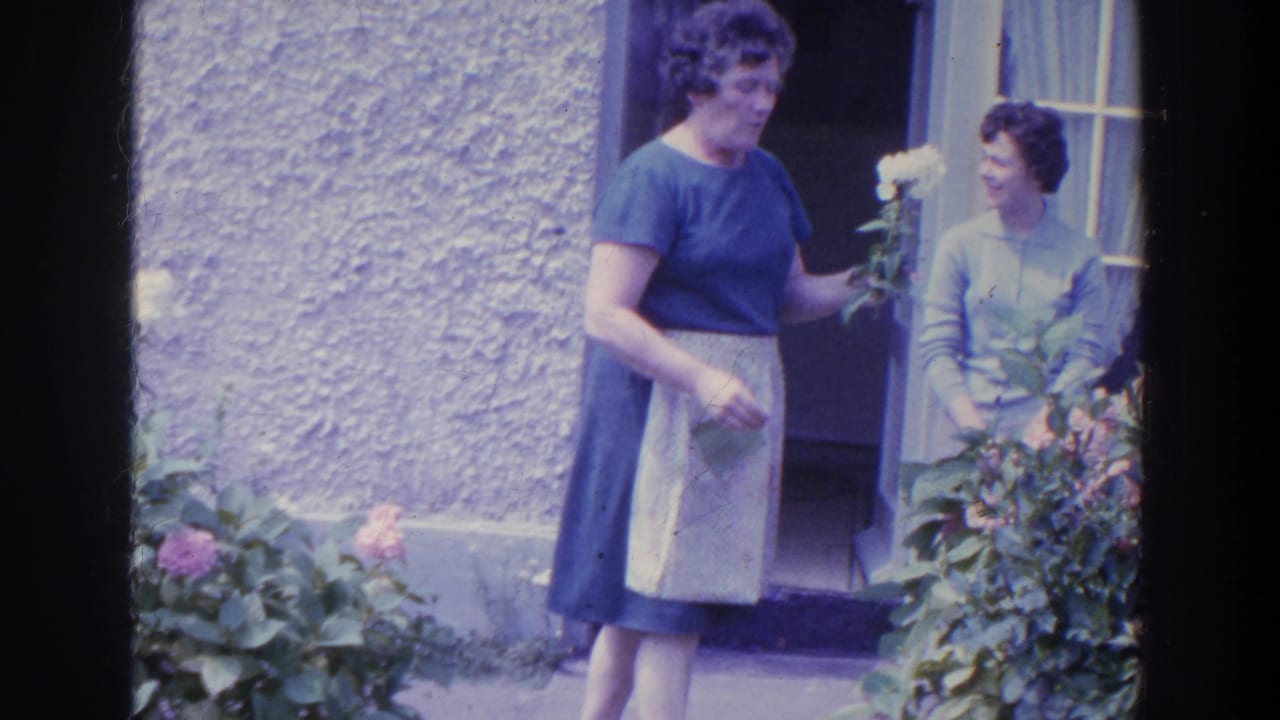 In 1961 Ireland, a woman in a blue dress and apron holds a white flower near garden bushes as another woman stands in the doorway.