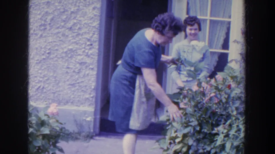 In 1961 Ireland, the woman in a blue dress and apron bends toward garden flowers while another woman smiles from the doorway.