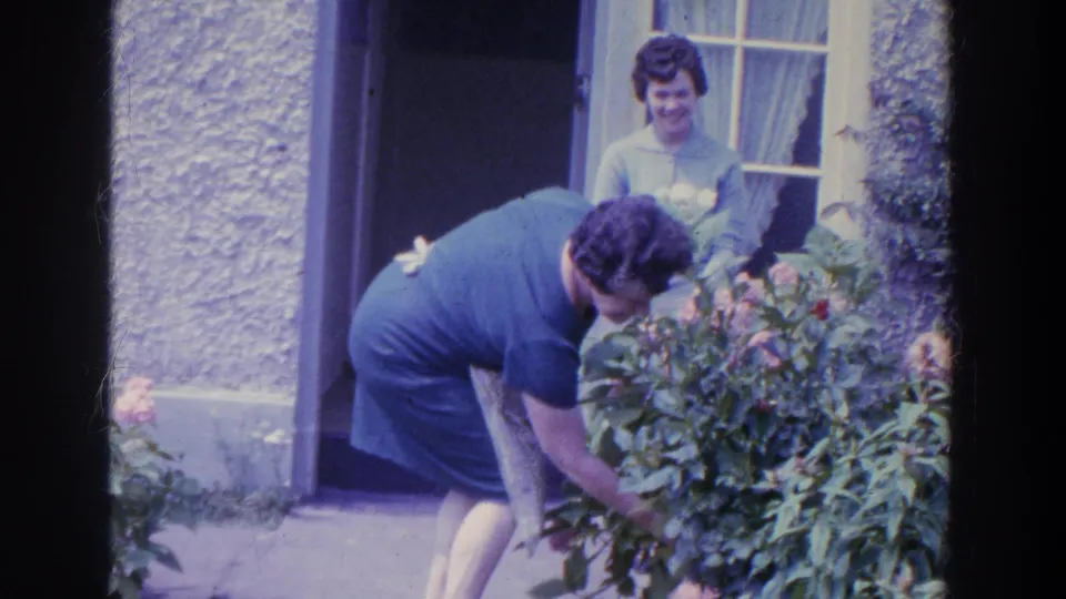 In 1961 Ireland, a woman in an apron reaches into a garden bush to pick flowers while another woman observes from the doorway.
