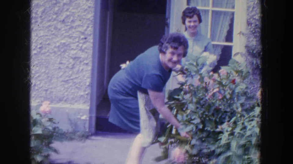 In 1961 Ireland, the woman smiles toward the camera while picking flowers beside the doorway, with another woman standing behind her.