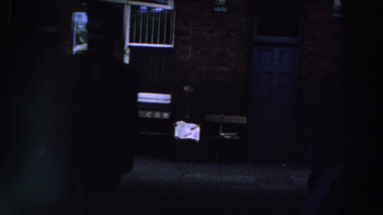 1961 Ireland 8mm frame showing people in deep shade near a brick wall, white-barred window, doorway, and small office sign.
