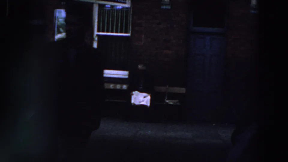 1961 Ireland 8mm frame showing people in deep shade near a brick wall, white-barred window, doorway, and small office sign.