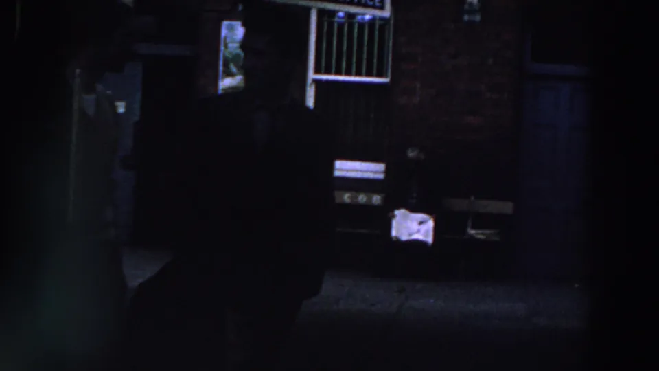 Low-light 1961 Ireland frame with a person partly visible in a doorway and shadowed figures crossing the foreground.