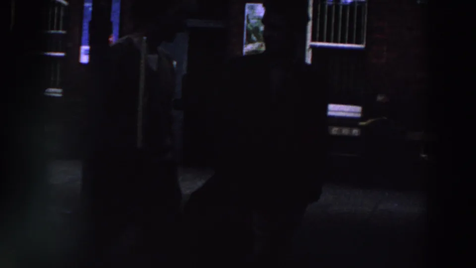 1961 Ireland 8mm frame showing several dark figures near a brick exterior, doorway, white-barred windows, and pavement.