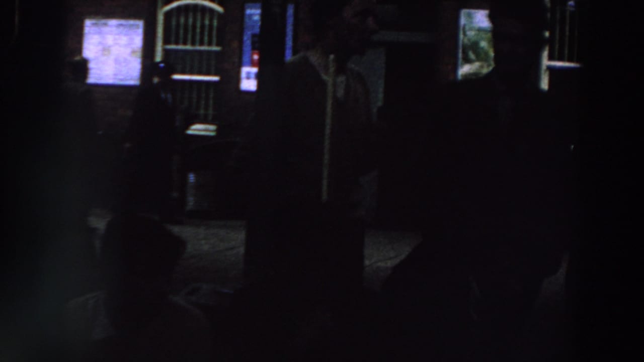 1961 Ireland home-movie frame showing shadowed figures near a brick exterior with lit notices and a barred arched doorway.