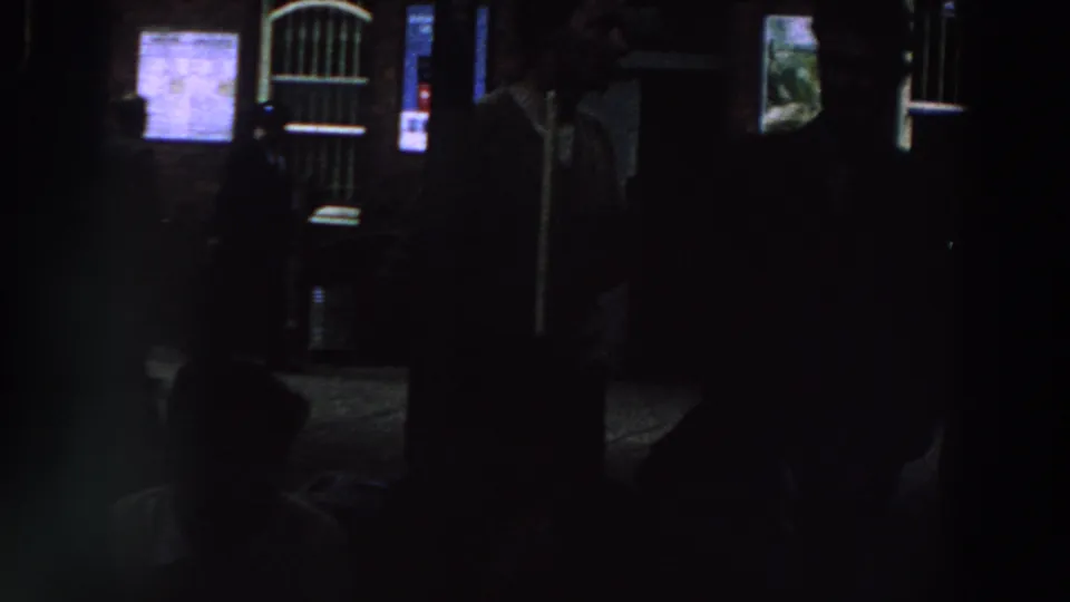1961 Ireland home-movie frame showing shadowed figures near a brick exterior with lit notices and a barred arched doorway.