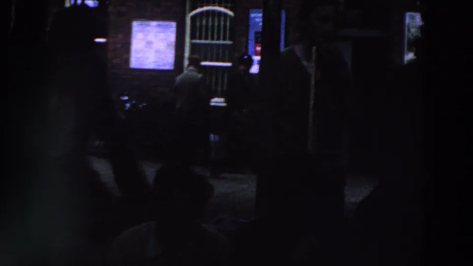 Low-light 1961 Ireland frame with indistinct people in front of a brick building, lit notices, and an arched barred doorway.