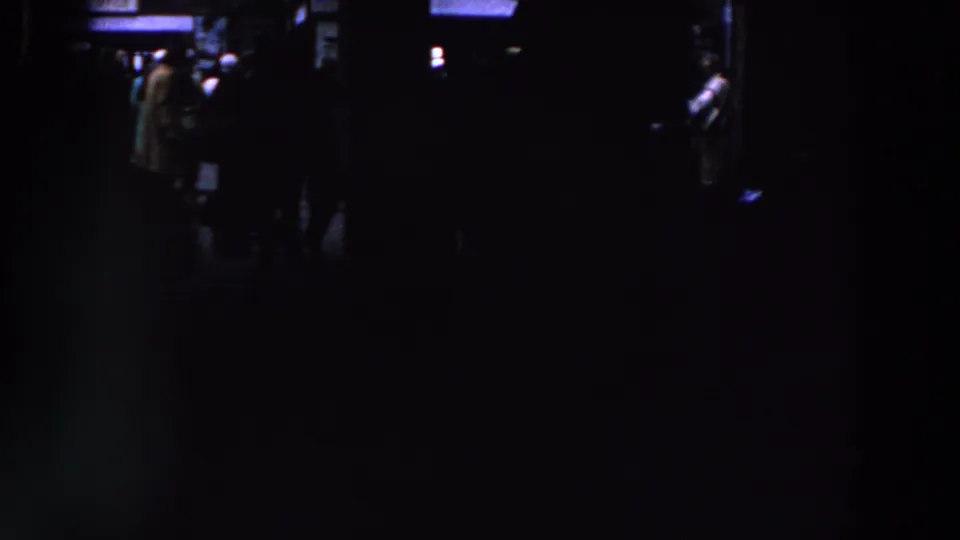 1961 Ireland home-movie frame with a shadowed indoor crowd and a few people briefly visible near blue-white light.