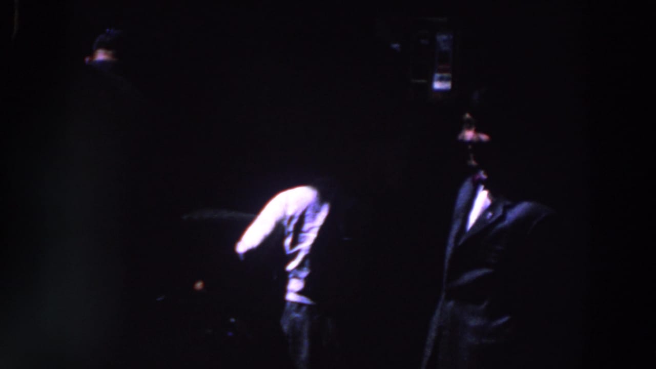 1961 Ireland home-movie frame showing a dark indoor gathering with suited figures, heavy underexposure and visible film grain.
