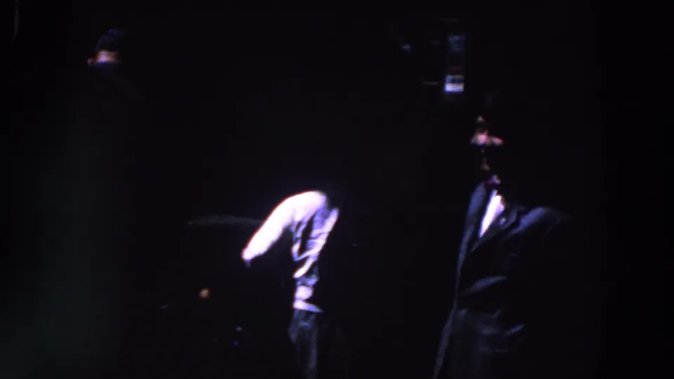 1961 Ireland home-movie frame showing a dark indoor gathering with suited figures, heavy underexposure and visible film grain.