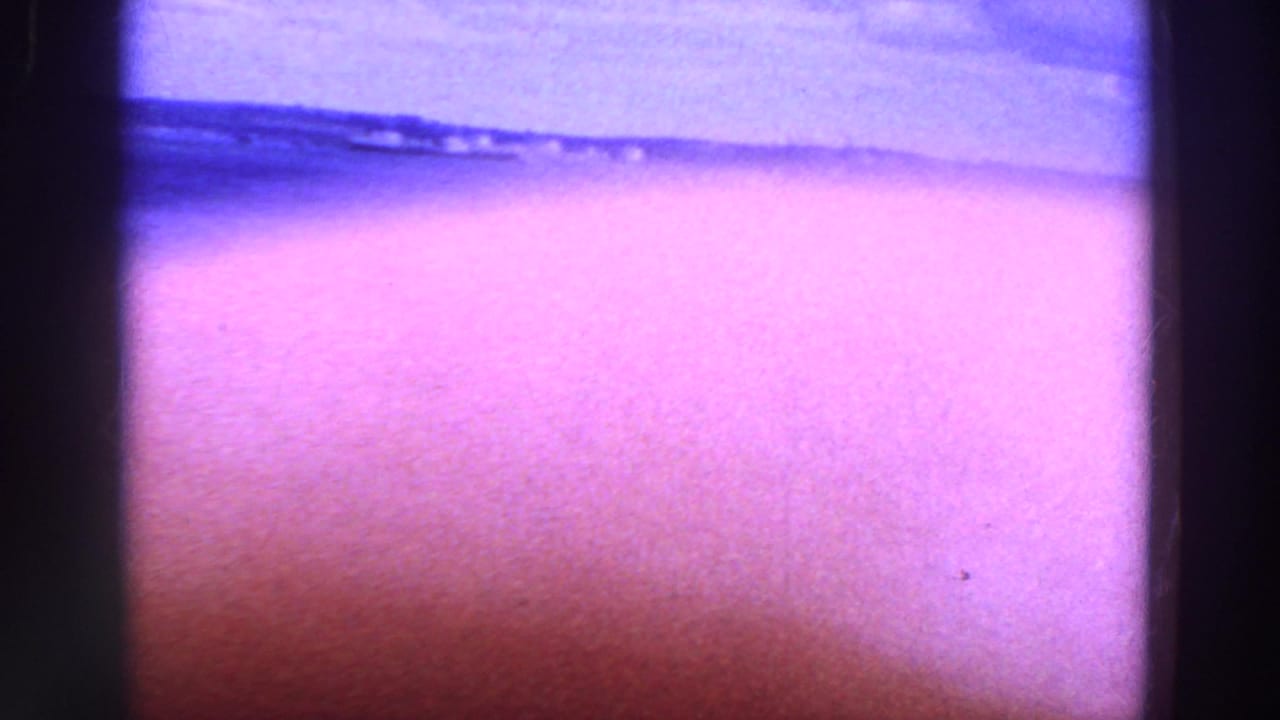 Purple-tinted beach view with water, distant shoreline and dark film-frame edges.