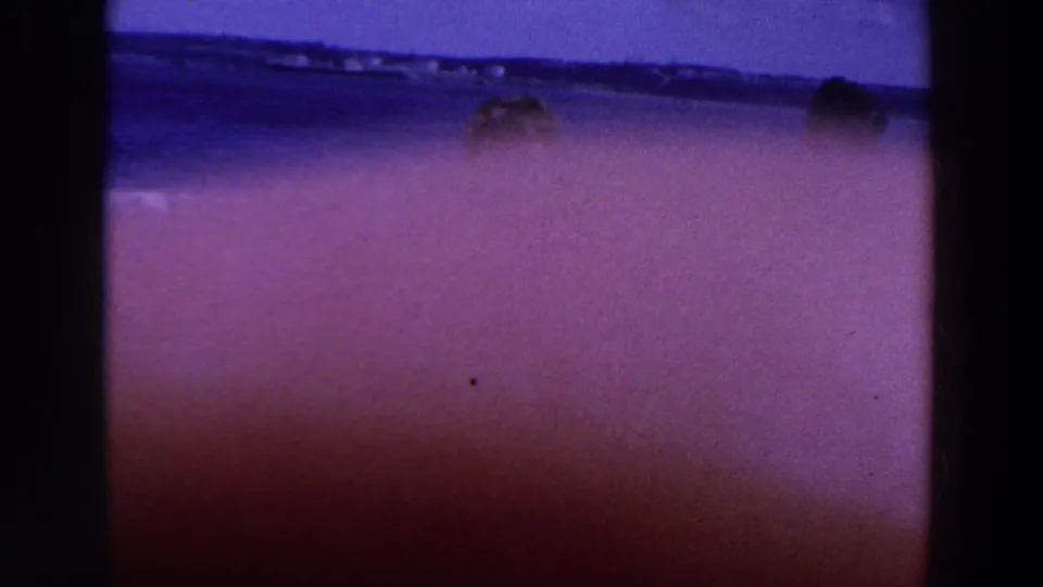 1961 Ireland home-movie frame showing partly obscured people wading at a beach with purple tint and grain.