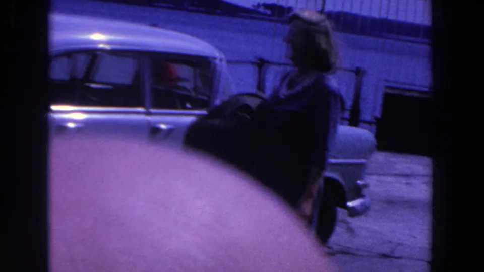 A handheld 1961 Ireland home-movie frame shows the woman moving near the side of a pale classic car on the pier.