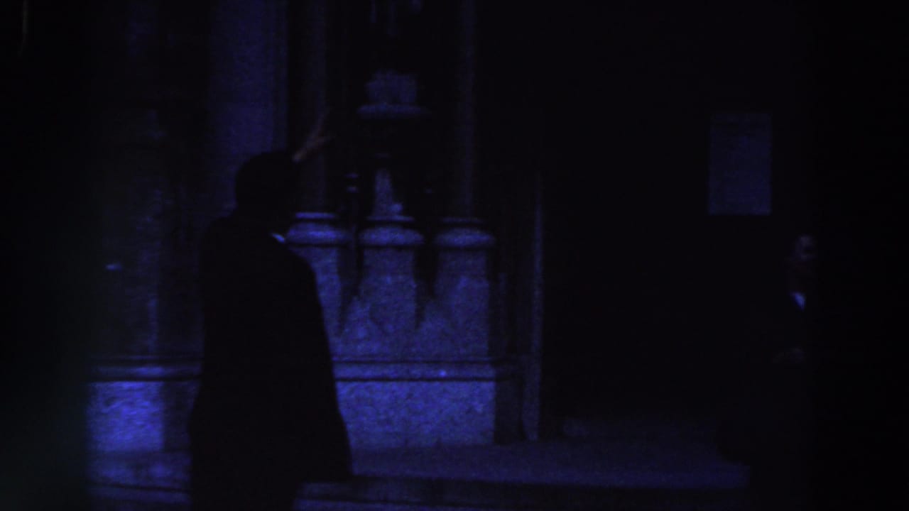 Silhouetted man points upward near a dim stone facade.